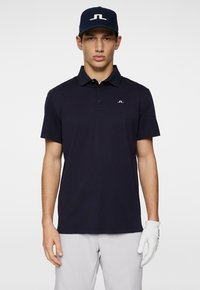 Marinblå polo skjorta med korta ärmar, krage och vitt logo, tillsammans med ljusgrå byxor och en vit golfhandske.