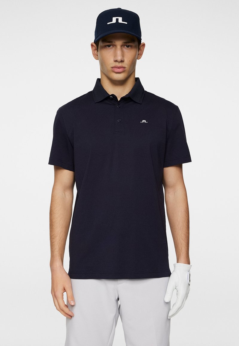 Marinblå polo skjorta med korta ärmar, krage och vitt logo, tillsammans med ljusgrå byxor och en vit golfhandske.