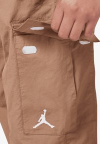 Bruine stoffen shorts met een gestructureerde afwerking, voorzien van een zijzak en een witte geborduurde Jumpman-logo. Klittenbandaccenten zijn zichtbaar.