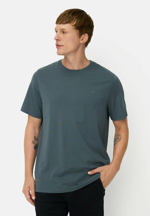 Ung mand med kort brunt hår iført en ensfarvet blågrå T-shirt og sorte bukser, der kigger til siden med den ene hånd i lommen.