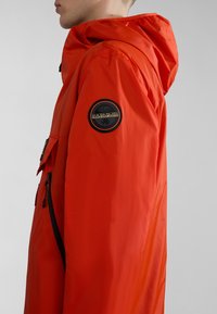 Zijaanzicht van een persoon die een feloranje hoodie-jack draagt met een zwart rond Napapijri-logo op de bovenarm.