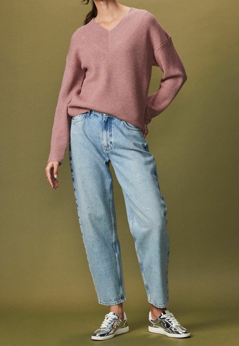 Femme portant un pull côtelé rose, un jean bleu clair avec de petits motifs floraux et des baskets argentées métalliques, debout devant un fond vert.