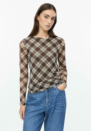 Femme aux longs cheveux bruns portant un haut à manches longues à carreaux marron et beige et un jean bleu, une main dans la poche.