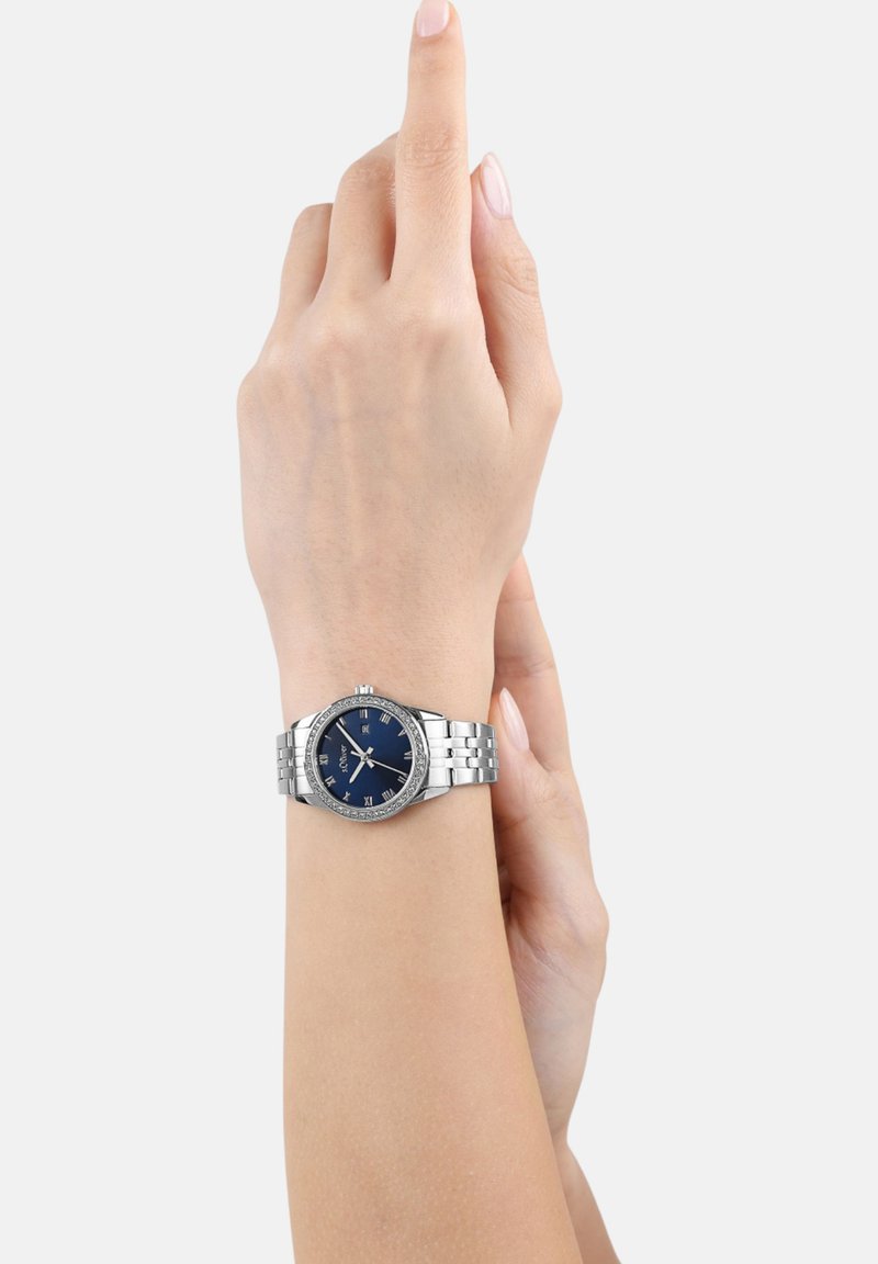 Silberner Edelstahl-Armbanduhr mit blauem Zifferblatt, römischen Ziffern und einem dekorativen Gehäuse. Am Handgelenk mit neutralem Hautton getragen.