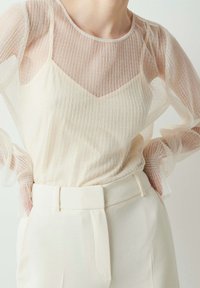 Przezroczysta bluzka z długim rękawem w pionowe paski w delikatnym odcieniu ecru, nałożona na jednolitą, jasnobeżową bazę, zestawiona z dopasowanymi, białymi spodniami z wysokim stanem.