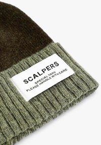 Gorro de punto verde con textura acanalada y una sección superior marrón oscuro. Incluye una etiqueta blanca con texto negro con instrucciones de cuidado.