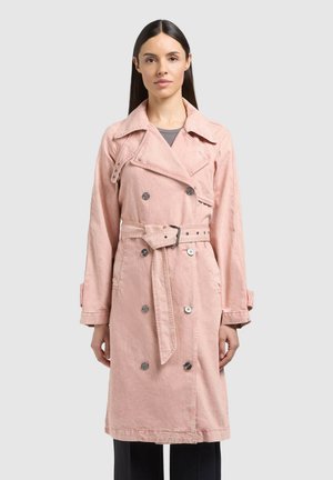 Žena s rovnými tmavými vlasmi má na sebe svetloružový dvojradový trenčkot s opaskom cez tmavé nohavice, stojí pred svetlosivým pozadím.