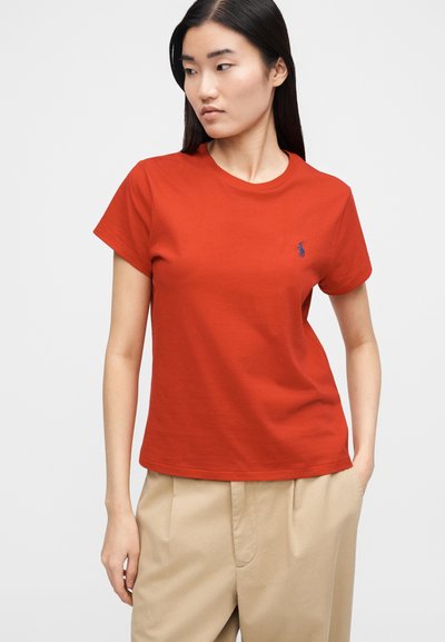 Piros pamut póló kerek nyakkivágással és rövid ujjal, a bal mellrészen egy kis kék hímzett logóval.