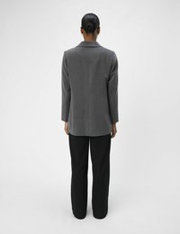 Blazer taillé gris avec une silhouette structurée et des manches longues, associé à un pantalon noir à jambe droite. Texture de tissu lisse, détails minimaux.