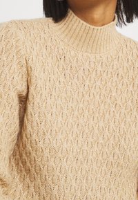 Nahaufnahme eines beigen, kabelgestrickten Pullovers mit einem gerippten hohen Kragen, getragen von einer Person mit kurzem dunklem Haar.