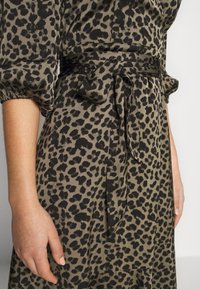Detailní záběr na ruku osoby na šatech s leopardím potiskem, které mají uvázaný pás a nabrané rukávy, na světlém pozadí.