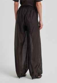 Žena v širokých, poloprůhledných černých kalhotách s elastickým pasem a stahovanými lemy, stojící před jednobarevným pozadím.