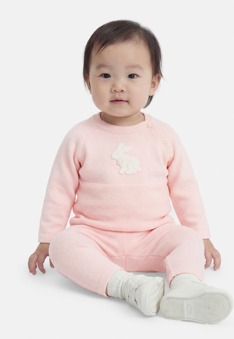 Νήπιο καθισμένο, φοράει ένα ροζ πλεκτό σύνολο με σχέδιο λευκού κουνελιού και λευκές κάλτσες και παπούτσια, με φόντο λευκό.