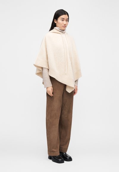Poncho en laine beige avec bords côtelés et capuche à cordon, superposé à un pull beige clair, associé à un pantalon marron à jambes larges et des chaussures noires.