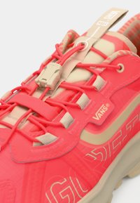 Bright coral and beige Vans sneaker close-up, showing elastic lace system, branded pull tab, and "Vans" logo on side and tongue.
