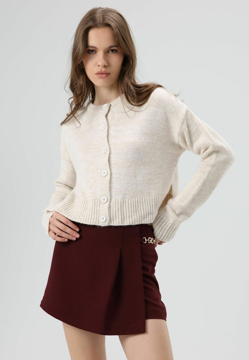 Cream knit cardigan with a round neckline and ribbed hem, featuring six white buttons. Paired with a maroon pleated mini skirt.