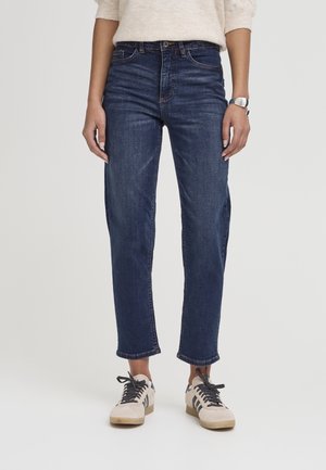 Femme portant un jean droit bleu foncé, un pull beige, une montre au poignet gauche et des baskets blanches avec des rayures noires et des lacets.