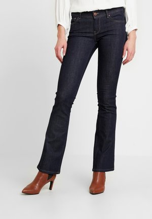 Kobieta ubrana w granatowe dżinsy typu bootcut i brązowe, szpiczaste botki na wysokim obcasie, stojąca na jednolitym tle.