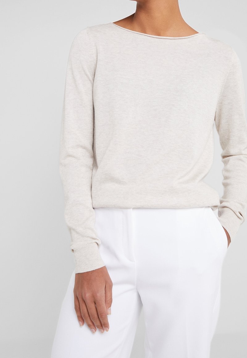 Un pull beige clair avec un large col, des manches longues et des poignets côtelés, associé à un pantalon blanc. Texture douce, design minimaliste.