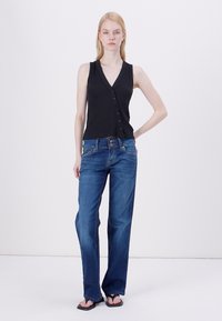 Černý top bez rukávů s detaily knoflíků, v kombinaci s modrými širokými džíny. Modelka stojí na neutrálním pozadí a má na sobě černé žabky.