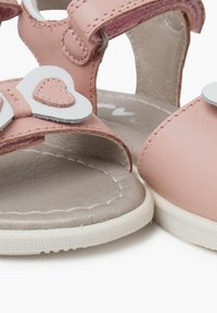 Sandalias sintéticas rosas con acentos blancos en forma de corazón, correas de gancho y bucle, y plantillas texturizadas grises. Cuenta con una suela acolchada y flexible.