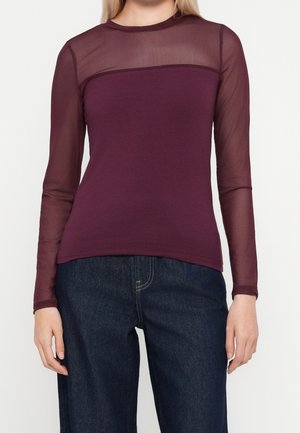 Vrouw draagt een aansluitende bordeauxrode top met lange mouwen en een transparant mesh bovenborst- en mouwgedeelte, gecombineerd met donkerblauwe jeans.
