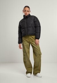 Svart pufferjakke med geometrisk mønster, høy krage og glidelås, kombinert med olivengrønne cargo-bukser og hvite sneakers.