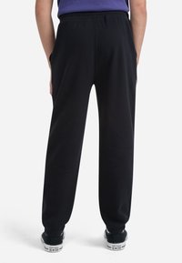 Pantalones de chándal negros con cintura elástica, bolsillos laterales, piernas ajustadas y puños acanalados. Llevados con zapatillas negras con detalles en blanco.