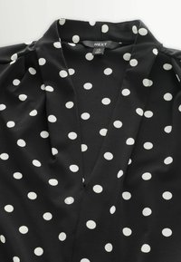 Fekete blúz fehér pöttyökkel, mély V-nyakkivágással és puha anyaggal, sima textúrát és folyékony, áramló kialakítást bemutatva.