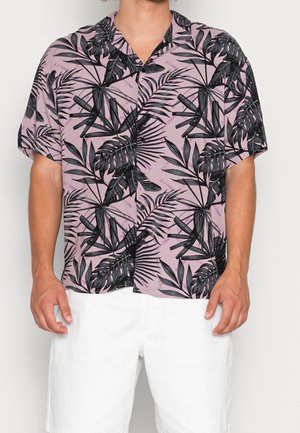 Homme portant une chemise à manches courtes boutonnée avec un imprimé de feuilles tropicales noires sur fond mauve et un short blanc.