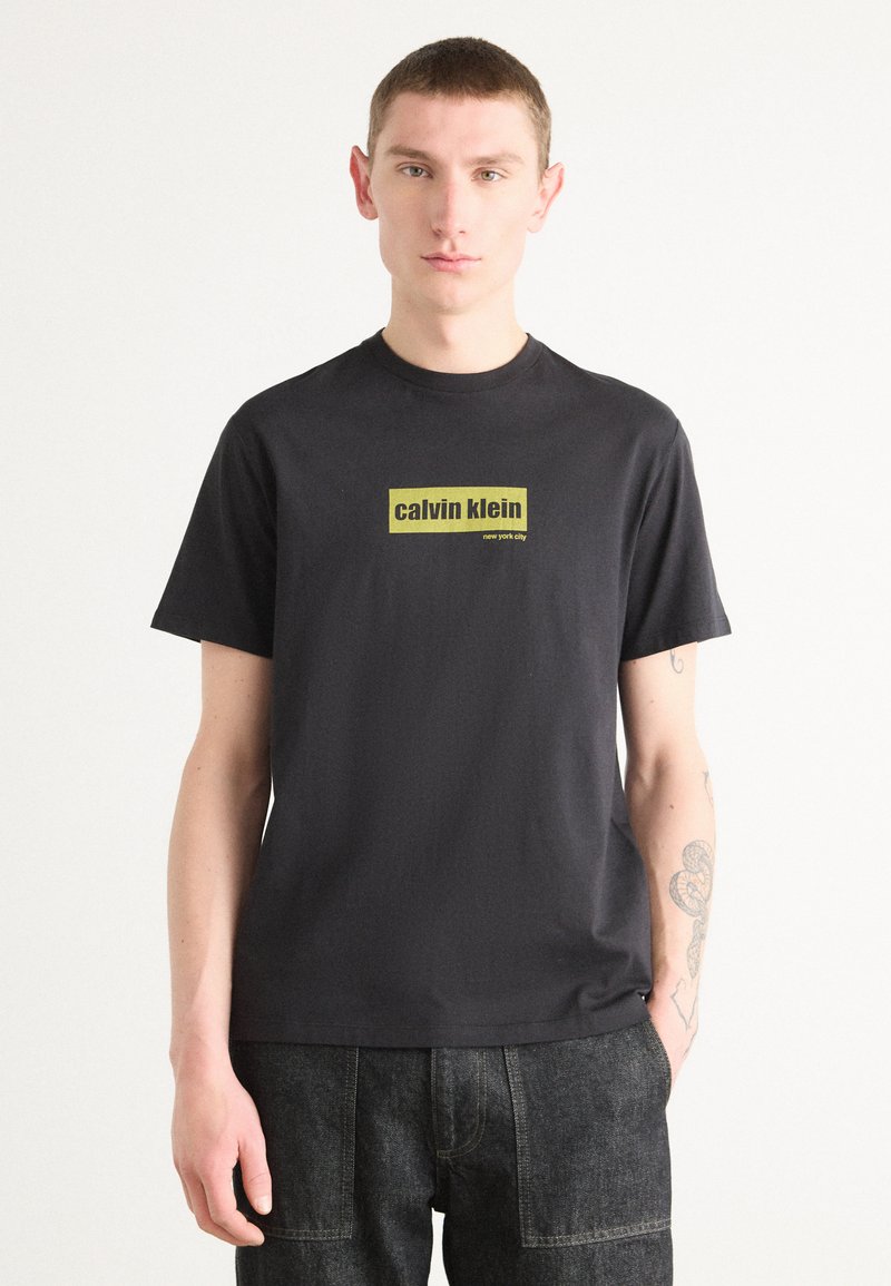 Svart bomull t-skjorte med et rektangulært gult logo som leser "calvin klein new york city" på forsiden. Korte ermer, rund hals.