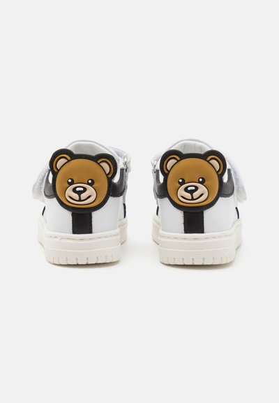 Zapatillas blancas con rayas negras y acentos de goma en forma de oso en el talón. Presenta superposiciones texturizadas y una suela acolchada.