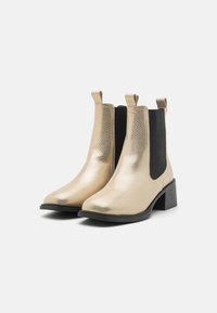 Bottes cheville dorées avec une finition texturée, panneaux élastiques noirs sur les côtés et talon bloc. Équipées de languettes pour un enfilage facile.