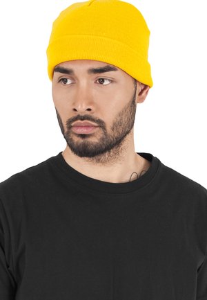 Yellow knit beanie with a ribbed texture worn with a black crewneck t-shirt, featuring a close-up of the hat and part of the model's face.