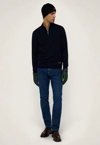 Námořnický pletený svetr se čtvrtinovým zipem, zelené rukavice, modré džíny a hnědé boty. Svetr má jemný vzor a na lemu hnědou štítek.