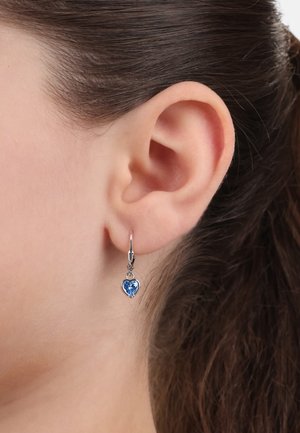 Silberne herzförmige Ohrringe mit einem blauen Edelstein, ausgestattet mit einem Klappverschluss, an einem Modell-Ohr präsentiert. Glatte und glänzende Oberfläche.