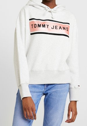 Osoba oblečená v světle šedé mikině s kapucí s nápisem "TOMMY JEANS" v růžové a černé, doplněná světle modrými džínami.