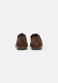 Chaussures en daim marron avec un petit talon, présentant un dos texturé et une doublure en tissu bleu visible à l'ouverture. Semelle en caoutchouc plate.