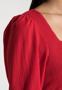 Röd texturerad blus med puffärmar och rundad halsringning, som framhäver ett zigzag-mönster och mjukt, vävt material.