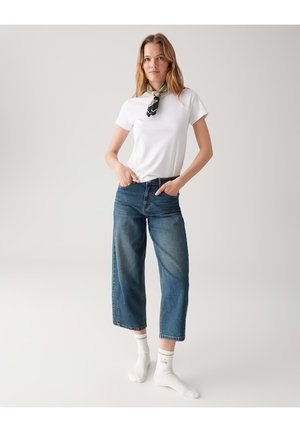 Weißes T-Shirt, hoch taillierte blaue Jeans mit weitem Bein, getragen mit weißen Socken. Ein grüner gemusterter Schal ist um den Hals gebunden.