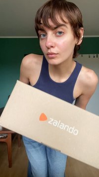 Caja de cartón marrón con el logo de "zalando", sostenida por una persona que lleva una camiseta sin mangas de canalé azul marino y jeans de mezclilla azul, en un espacio interior luminoso.
