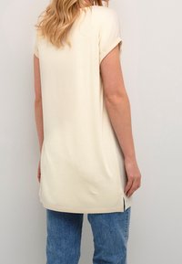 Tunique couleur crème avec des manches retroussées, fabriquée en tissu doux. Présente des fentes sur les côtés au niveau de l'ourlet et une coupe décontractée. Associée à un jean bleu clair.