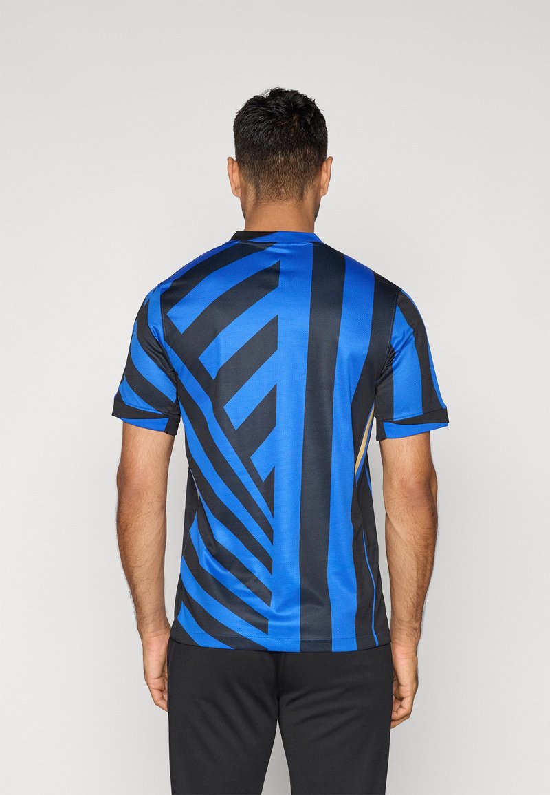 Hombre de pie de espaldas, vistiendo una camisa de manga corta azul y negra con llamativas rayas diagonales y verticales, combinada con pantalones negros.
