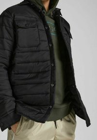 Chaqueta acolchada negra con cierre frontal, dos bolsillos de pecho con botones y un acabado liso, utilizada sobre una sudadera verde y pantalones crema.