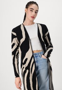 Adīts kardigāns ar melni-beige zebras rakstu, atvērta priekšpuse, garas piedurknes un teksturēts audums. Vilkts virs baltas blūzes un džinsiem.