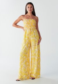 Combinaison florale jaune avec un corsage smocké sans bretelles et des jambes larges. Tissu léger et transparent avec une texture douce et fluide. Sandales noires.