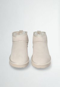 Un paio di pantofole in suede beige per l'inverno, con morbido rivestimento in pelliccia e suole in gomma, posizionate frontalmente su uno sfondo semplice.