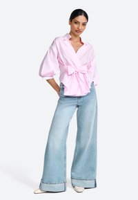 Blusa in cotone rosa con dettaglio di fiocco frontale, abbinata a jeans a gamba larga blu chiaro con orlo grezzo e accenti di cucitura delicati.