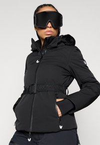 Μαύρο σακάκι σκι με φερμουάρ μπροστά, κουκούλα με γούνα και υφή υφάσματος. Διαθέτει αντίθετη μαύρη ζώνη και τσέ�πες με φερμουάρ.