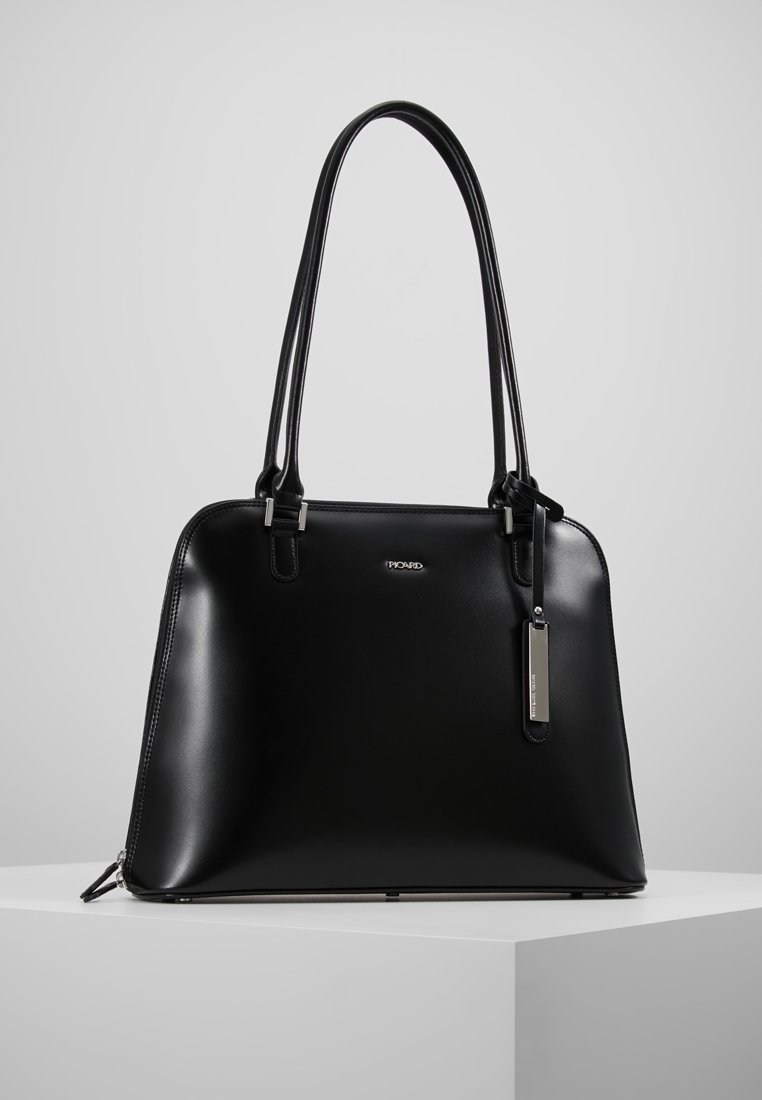 Sac à main en cuir noir avec deux longues lanières, logo de marque argenté et une étiquette métallique rectangulaire suspendue, présenté sur un piédestal blanc.